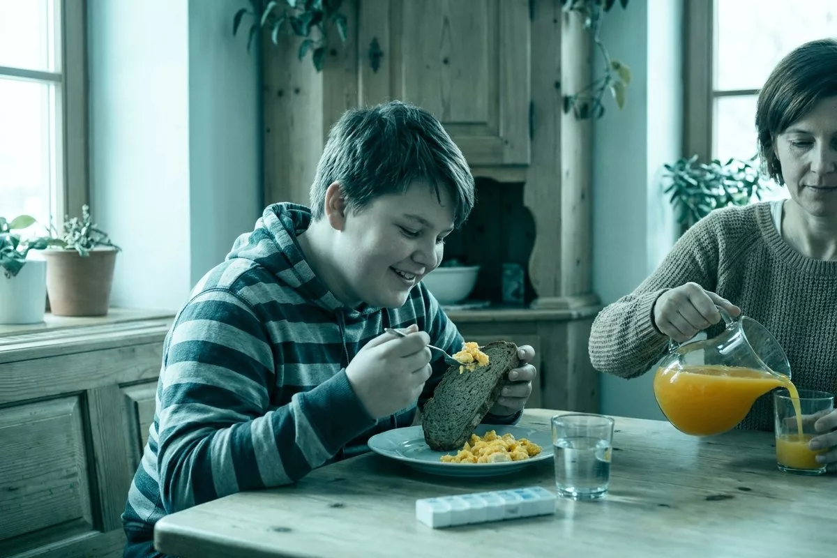 Ein Teenager isst ein herzhaftes Frühstück am Morgen, daneben ein Glas Wasser und eine Tablette