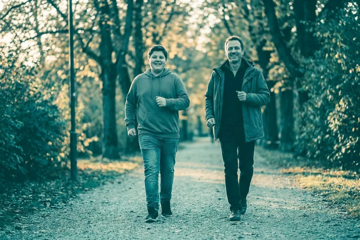 Ein Teenager geht energiegeladen mit einem Elternteil im Park spazieren, beide lächeln