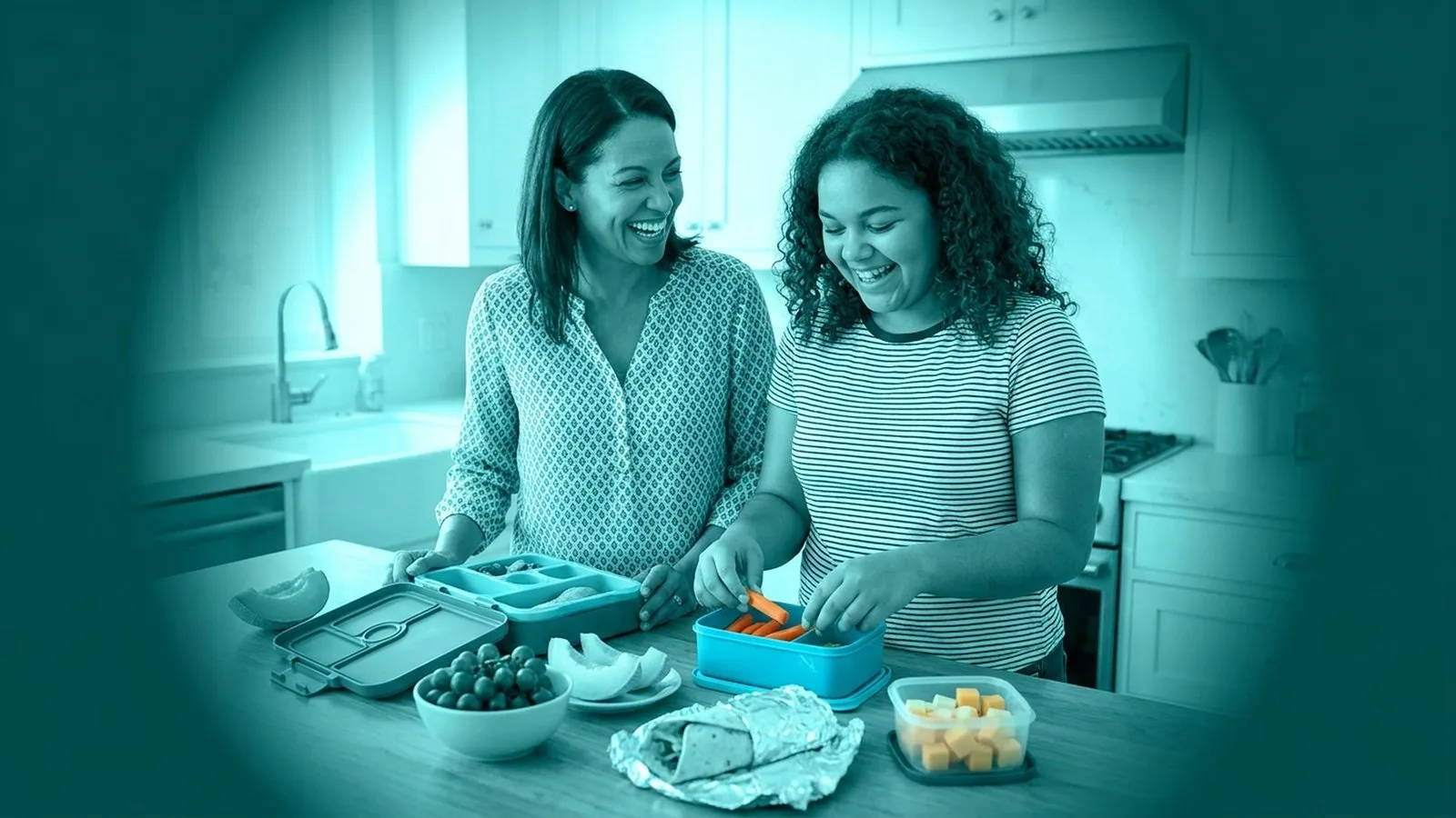 Mutter und Tochter befüllen gemeinsam lachend eine bunte Jausenbox mit frischem Obst und einem Wrap am Küchentisch